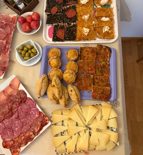 Mesa preparada para un evento con cocas de verdura, panades, cocarrois, queso y embutido en bandejas.